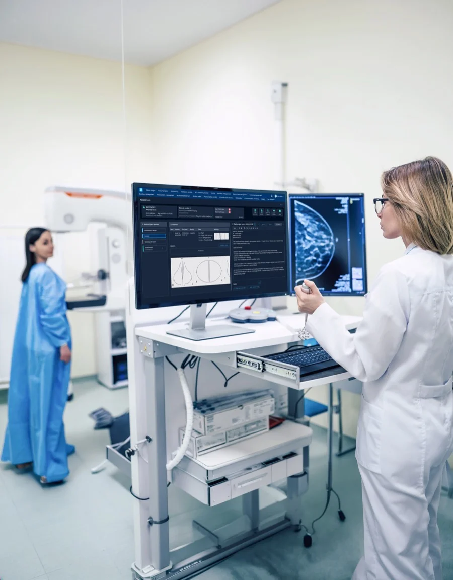 A radiologist reviews a mammogram and the structured Pinga assessment side by side on one workstation, while the patient waits at the mammography unit behind her